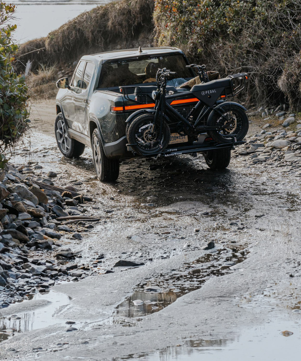 Two Pedal electric bikes mounted on a MotoTote Mini Dual electric bike carrier attached to a Rivian while traveling on a dirt road to the beach