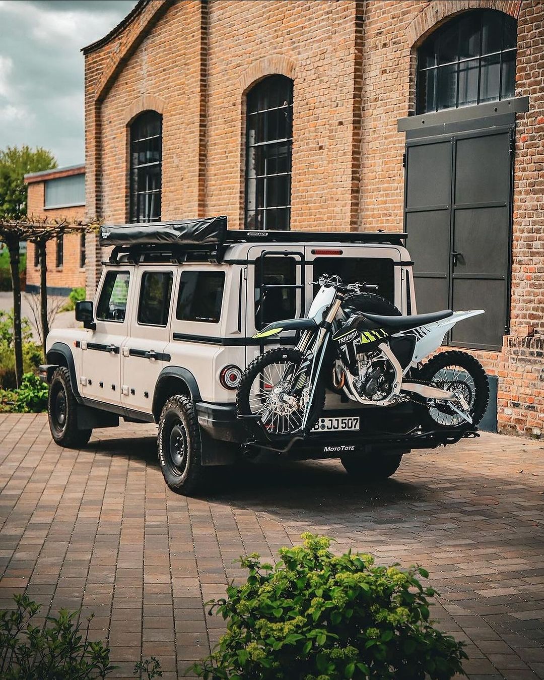 Triumph dirt bike mounted on a MotoTote Max+ motorcycle hitch carrier attached to a Land Rover Defender, showcasing premium motorcycle transport for overland and adventure travel.