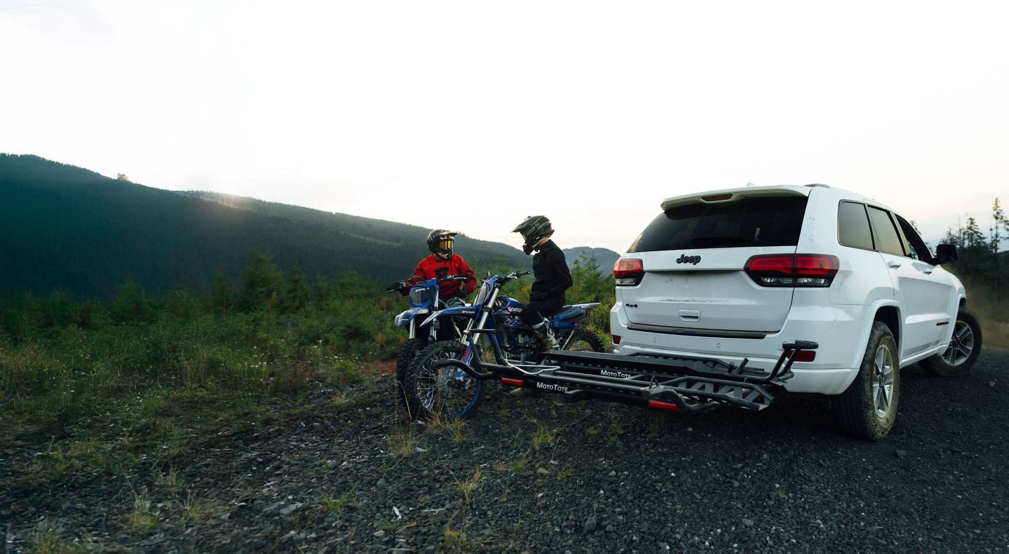 Two riders standing beside their dirt bikes mounted on a MotoTote Max Dual motorcycle hitch carrier, preparing for an off-road riding adventure in the outdoors.