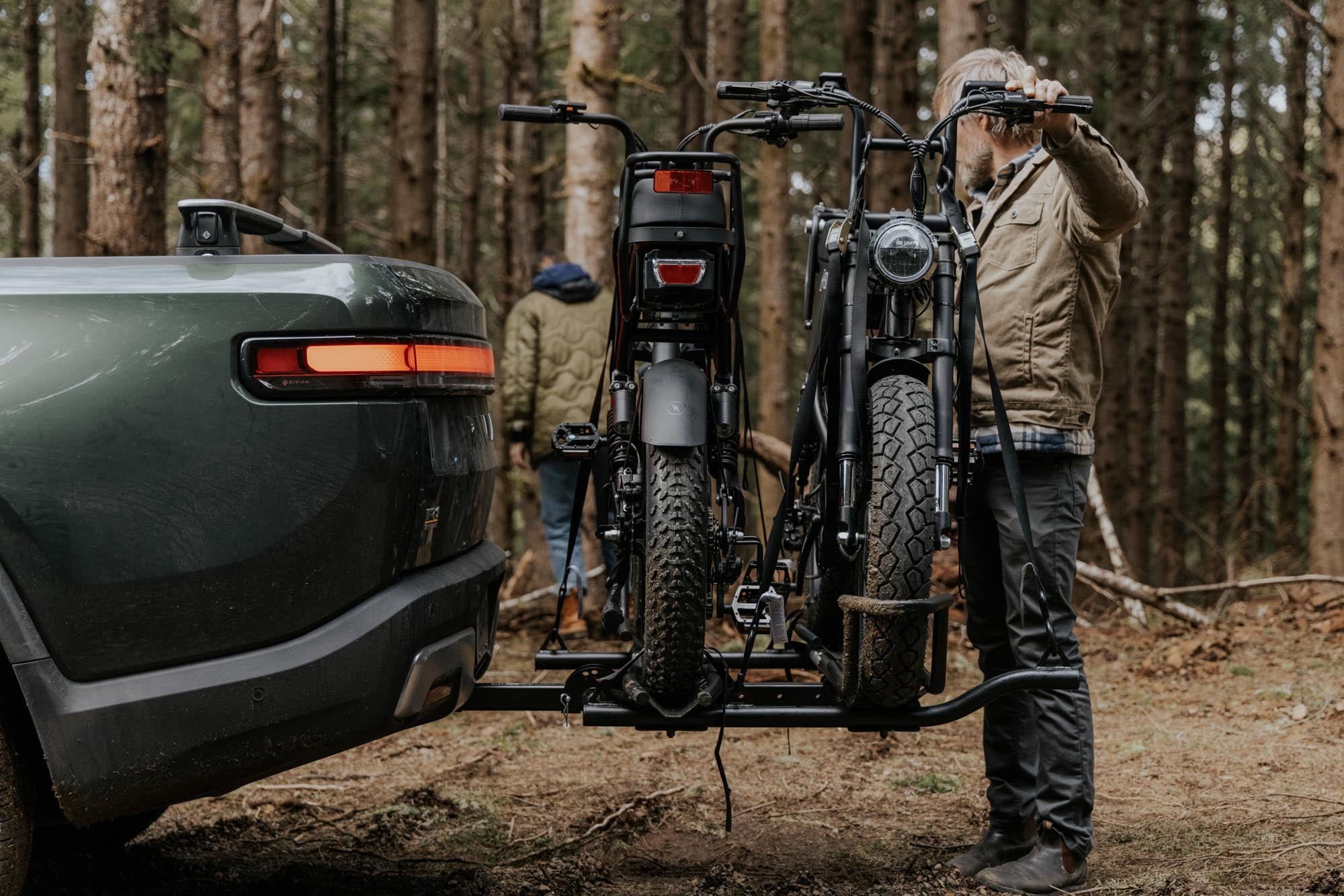 Pedal Electric e-bikes mounted on a MotoTote Mini Dual electric bike carrier attached to a Rivian truck in a forest campsite, showcasing secure transport for heavy electric bikes and outdoor adventure riding.