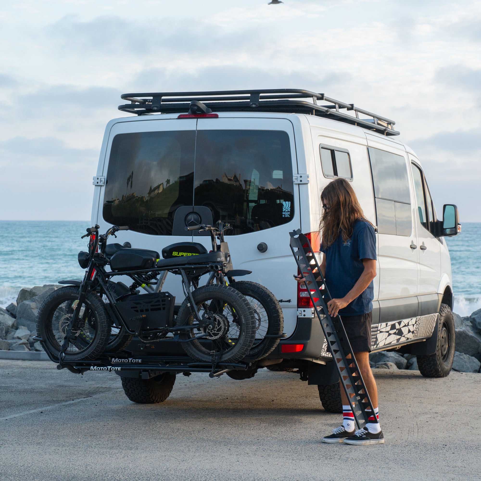 MotoTote Mini Dual electric bike carrier with ramp hauling two Super73 electric bikes behind a Mercedes Sprinter van by the coast, showcasing secure transport for heavy e-bikes and adventure travel.