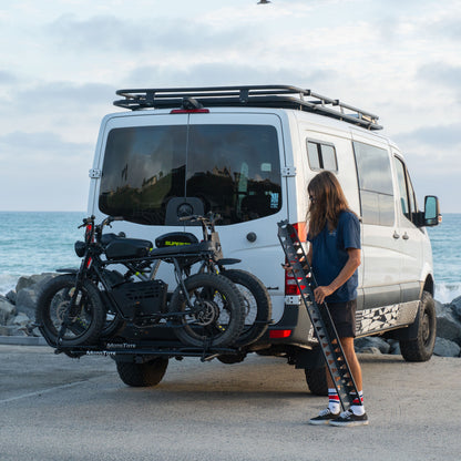 MotoTote Mini Dual electric bike carrier with ramp hauling two Super73 electric bikes behind a Mercedes Sprinter van by the coast, showcasing secure transport for heavy e-bikes and adventure travel.